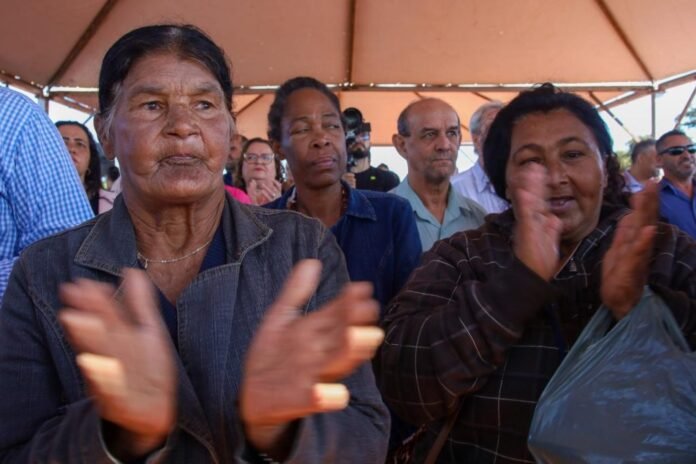 Group of people under a tent clapping, with two elderly women in the foreground wearing denim and plaid jackets.