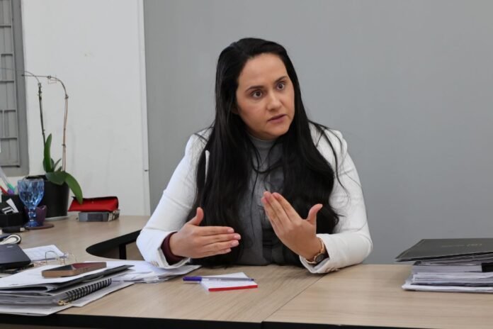 Woman in a white blazer explaining something, seated at a desk with papers and a plant nearby in an office.