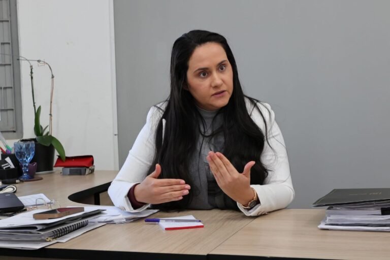 Woman in a white blazer explaining something, seated at a desk with papers and a plant nearby in an office.