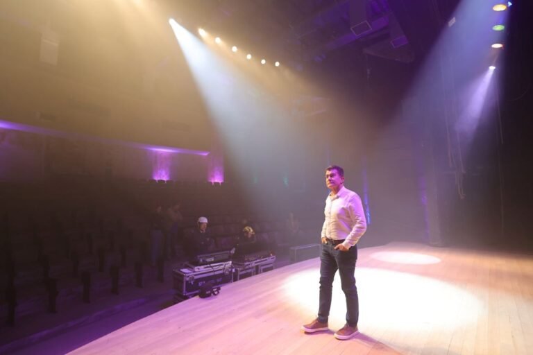 A man stands on a lit stage in a white shirt and jeans, hands in pockets, facing an empty audience hall with purple and warm spotlights.