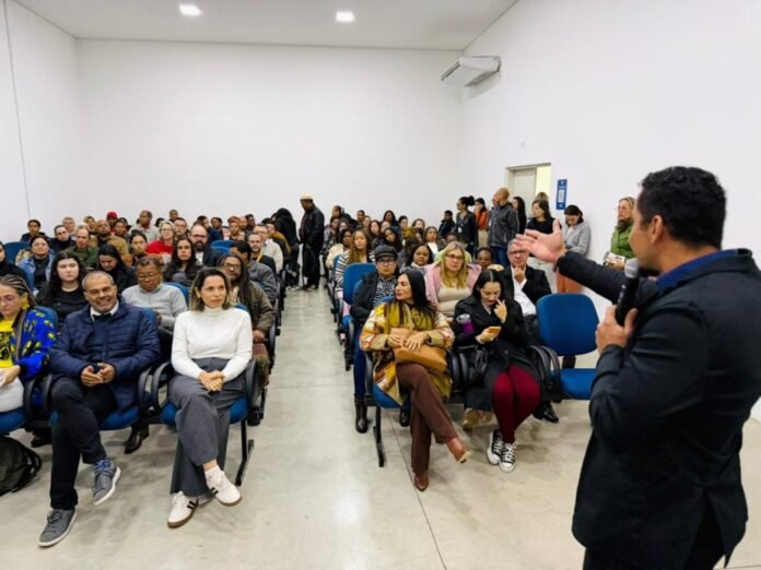 Speaker with a microphone addressing a diverse audience in a lecture room with rows of blue chairs and attendees listening attentively.