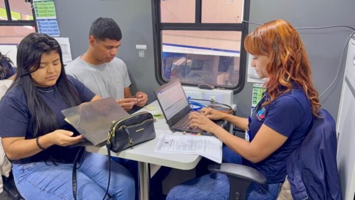 Team of three working at a small table inside a bus-like mobile office; woman with red hair types on a laptop while two colleagues review documents.