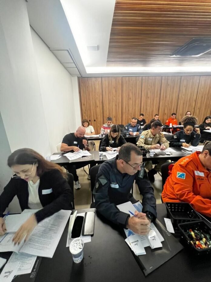 Group of people in a training room sitting at tables, some in uniforms, writing on papers and taking notes.