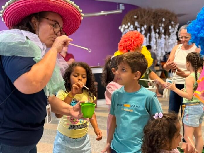 Adult in a bright pink hat blows bubbles for kids at a colorful indoor party, children watching and stowing bubble wands nearby.
