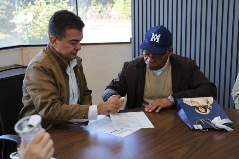Two men sit at a wooden table signing documents together in a bright office, with a blue bag on the right and a cup held by another person in the foreground nearby.