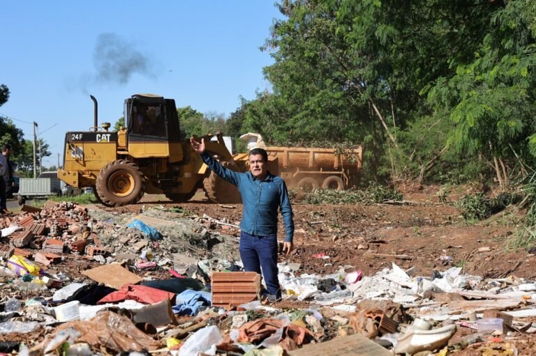 Man in blue shirt standing amid a junkyard full of scattered trash, pointing or gesturing toward the camera with a yellow bulldozer in the background.