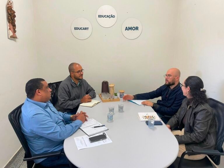 Four people sit around a small conference table in a bright room, engaged in a discussion with notebooks and papers on the table and cups nearby, wall art behind them reads Educação, Educart, Amor with three circles from left to right.
