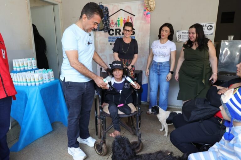 Man hands a bottle to a senior woman in a wheelchair while four adults stand nearby at a community center event with a dog nearby.