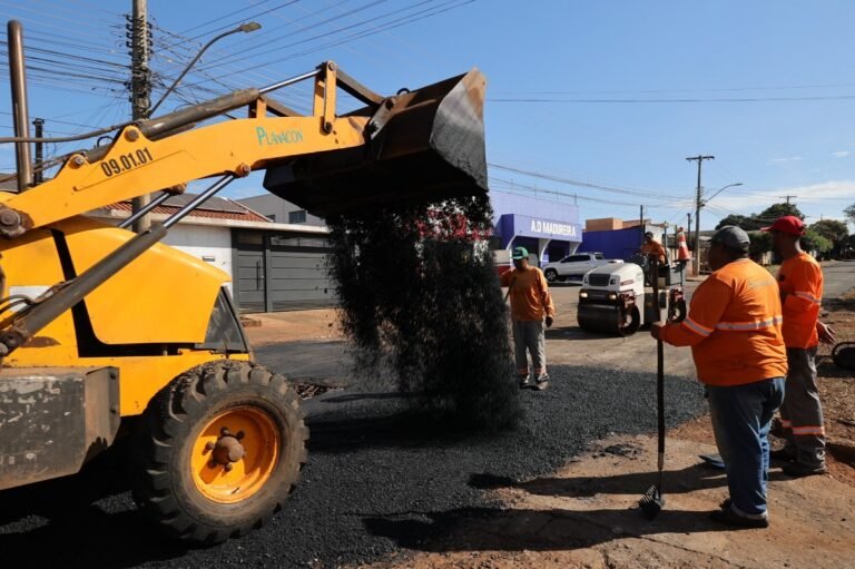 Yellow site excavator dumps hot asphalt onto a road as workers in orange vests monitor the paving process.