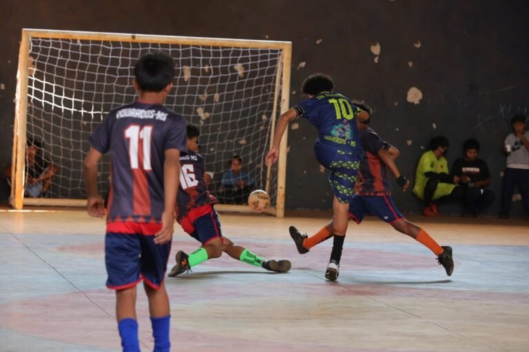 Futsal players chase the ball near the goal, with one player in blue number 10 jumping as others defend. Audience sits in the background on the right.