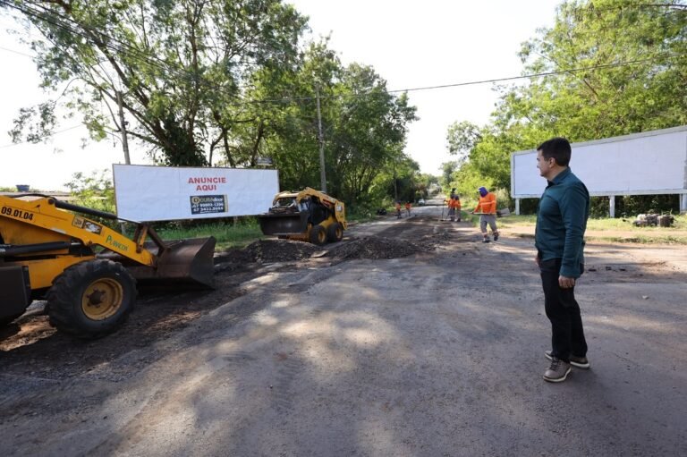 Road repair scene with two yellow construction machines, piles of asphalt, and workers in orange vests; man in teal shirt watches from the right.