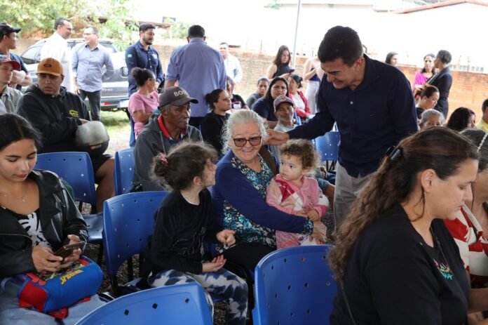 Man in a navy shirt greets or talks with an elderly woman holding a young child at an outdoor community gathering.