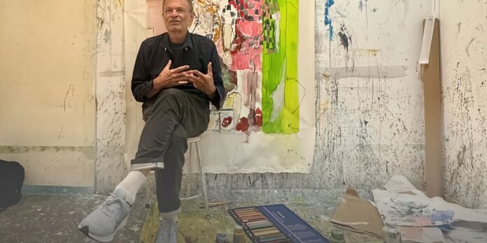 Man seated on a stool in a messy artist's studio, gesturing with his hands as he speaks; a large abstract painting with bright colors is behind him.