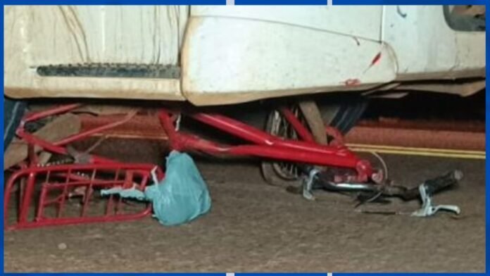 Red bicycle frame wedged under a white vehicle, with a blue bag on the ground and scattered tools nearby.