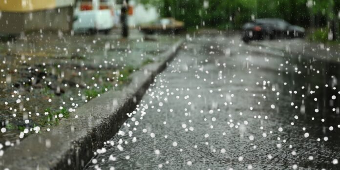 Stormy street scene with heavy hail bouncing off a wet curb and pavement, car and greenery in the background.