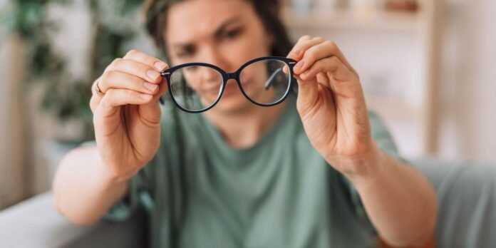 Person holds up a pair of black-rimmed glasses toward the camera, with a blurred background behind them.