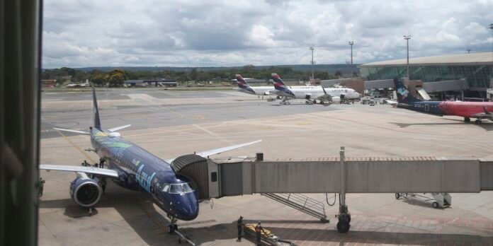 Blue commercial jet at a gate connected to a jet bridge, ground crew nearby on the tarmac with other planes visible in the background.