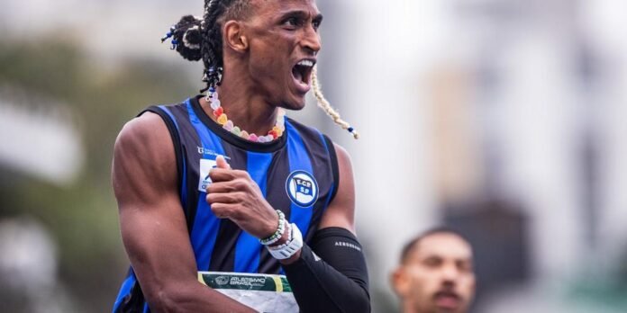 Male sprinter in a blue-and-black striped jersey shouting in celebration during a race, wearing a colorful beaded necklace and wristbands.