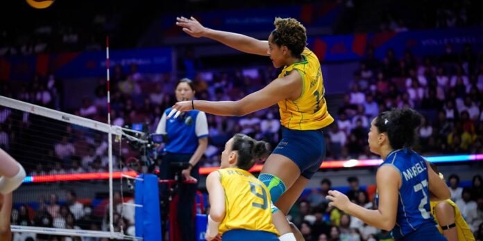 Female volleyball player in yellow jumps over the net during a match, teammates nearby in blue and yellow uniforms.