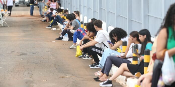 Group of diverse young people sitting along a sidewalk beside a tall blue fence, waiting in a long line.