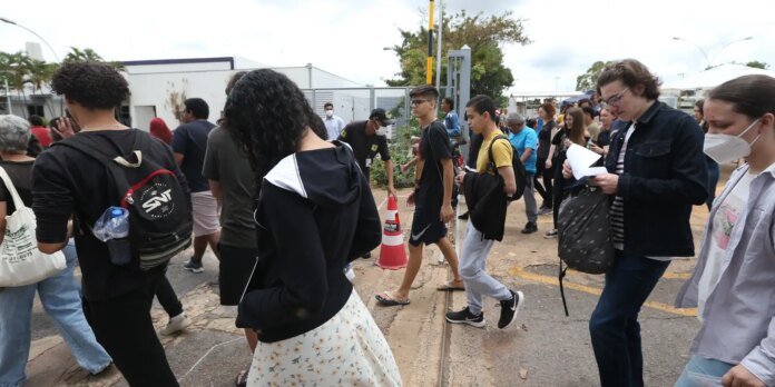 Group of diverse people walking along a paved street outdoors, some wearing backpacks and face masks, with a traffic cone nearby.