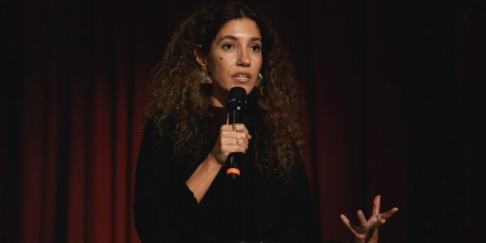 Woman with curly hair speaking into a handheld microphone on a dark stage.