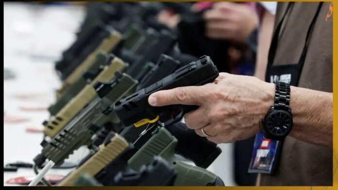 Close-up of a person at a gun show handling a black handgun from a row of pistols on display, with a wristwatch and badge visible on their wrist/vest.