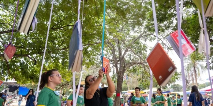 Children in green shirts participate in an outdoor book swap under a tree, with colorful books hanging from ribbons and an adult reaching up to place a book.