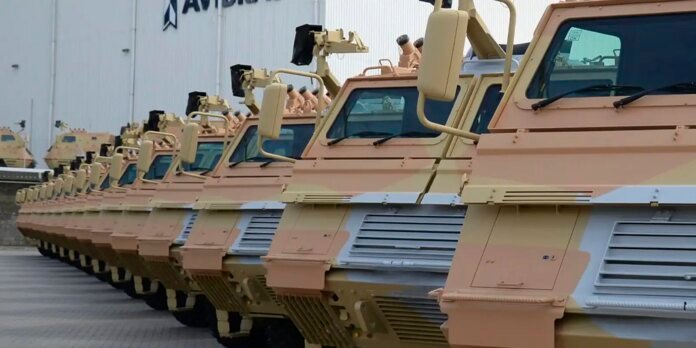 Row of desert-colored armored vehicles lined up outside a warehouse or hangar yard.