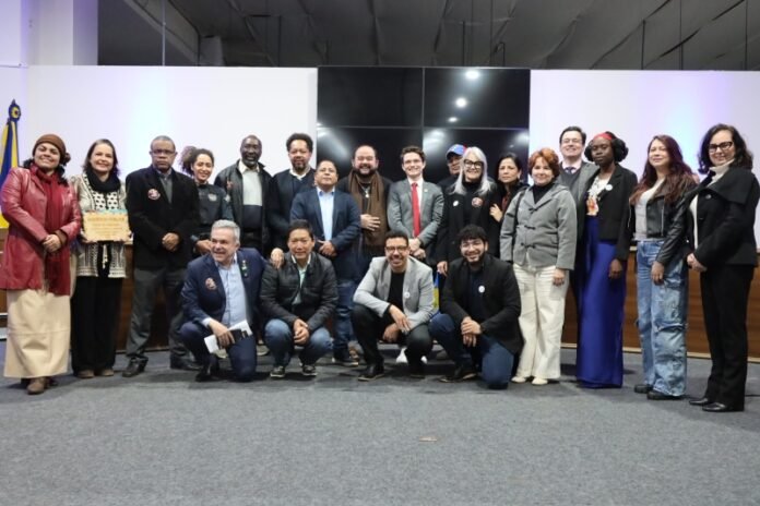 Diverse group of professionals posing for a group photo at a formal event on a conference stage, with a flag to the left and a large screen behind them.