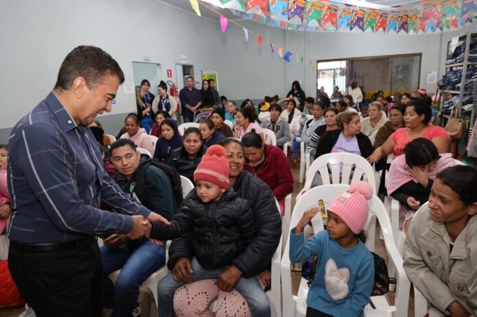Man in blue shirt shakes hands with a child in a warm jacket at a crowded community event hall.