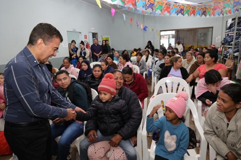 Man in blue shirt shakes hands with a child in a warm jacket at a crowded community event hall.