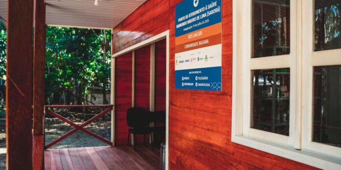 Red wooden clinic hut with a blue/orange signboard advertising health services, outdoors among green trees, with a window on the right side and a chair inside.