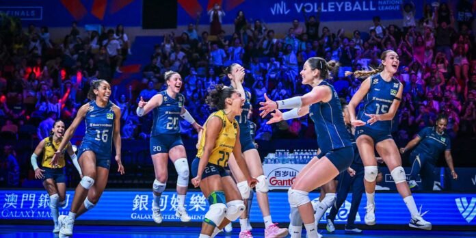 Female volleyball players in blue uniforms leap and celebrate on the court as teammates cheer and fans applaud in a blue-lit arena.