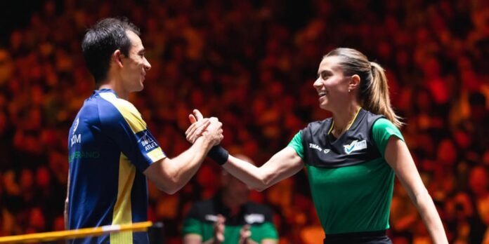 Two badminton players shake hands at the net after a match, smiling in a crowded arena; one in blue and yellow, the other in green and black.