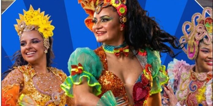 Three women in bright carnival costumes dancing and smiling on a blue backdrop stage.