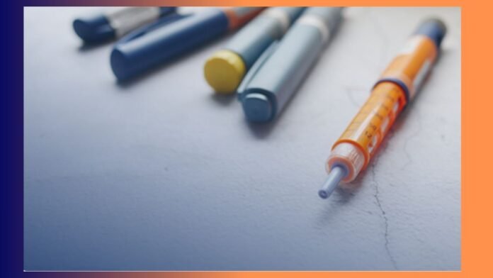 Close-up of a syringe with an orange plunger lying on a pale surface, with blurred colorful marker caps in the background.