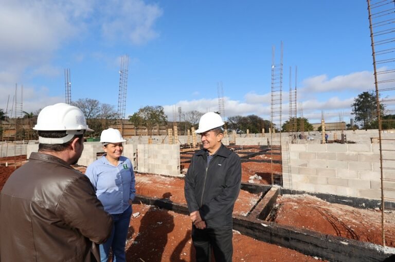 Three people wearing white hard hats talk on a construction site with partially built concrete walls and exposed rebar under a blue sky.