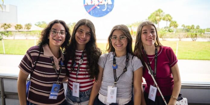Four smiling teenage girls with badges pose together outdoors with a NASA sign in the background at a sunny event.