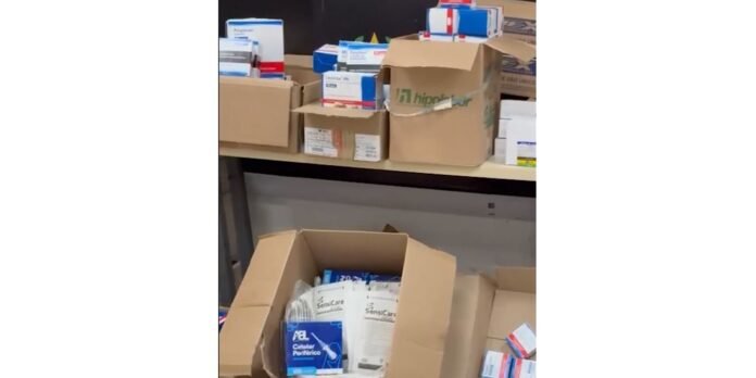 Open cardboard box on a table filled with assorted medical supplies and pill boxes in a storage area.