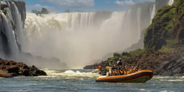 Orange inflatable tour boat carrying passengers on a choppy river beside a tall, misty waterfall.