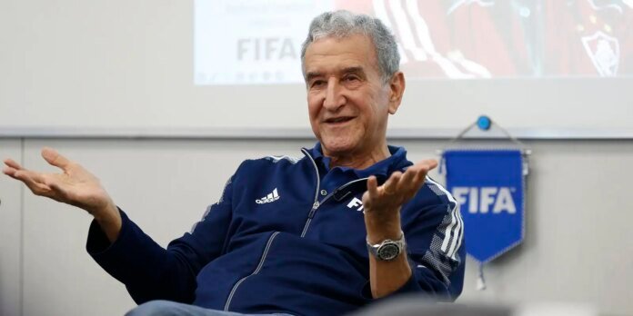 Smiling older man in a navy Adidas jacket speaks with open hands; a FIFA banner is in the background.