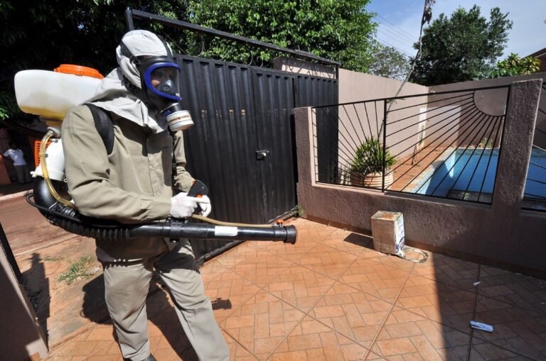 Pest control worker in full protective suit with respirator using a backpack sprayer to spray a patio near a pool.