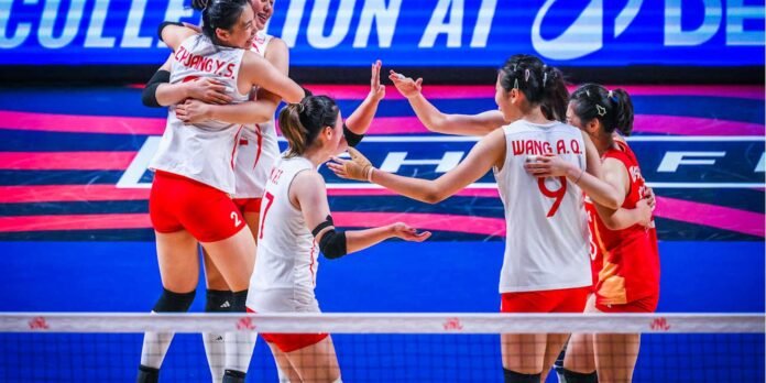 Group of female volleyball players in white and red uniforms celebrate at the net after a point on a blue court, arms raised in celebration.
