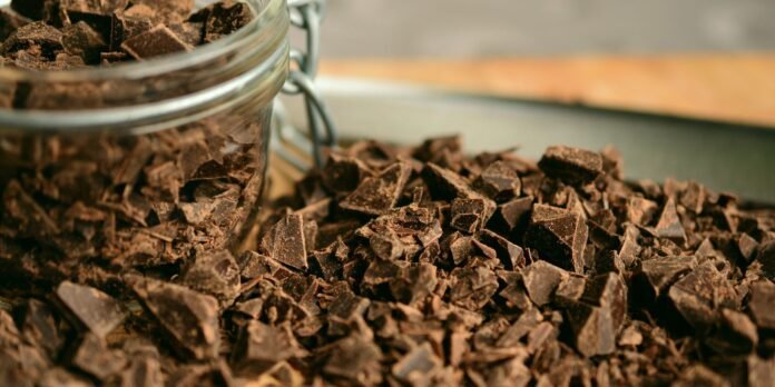 Chunks of dark chocolate spilling from a glass jar onto a wooden surface.