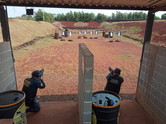Two officers in a shooting range crouch behind barrels, aiming at paper targets downrange under a covered shelter.