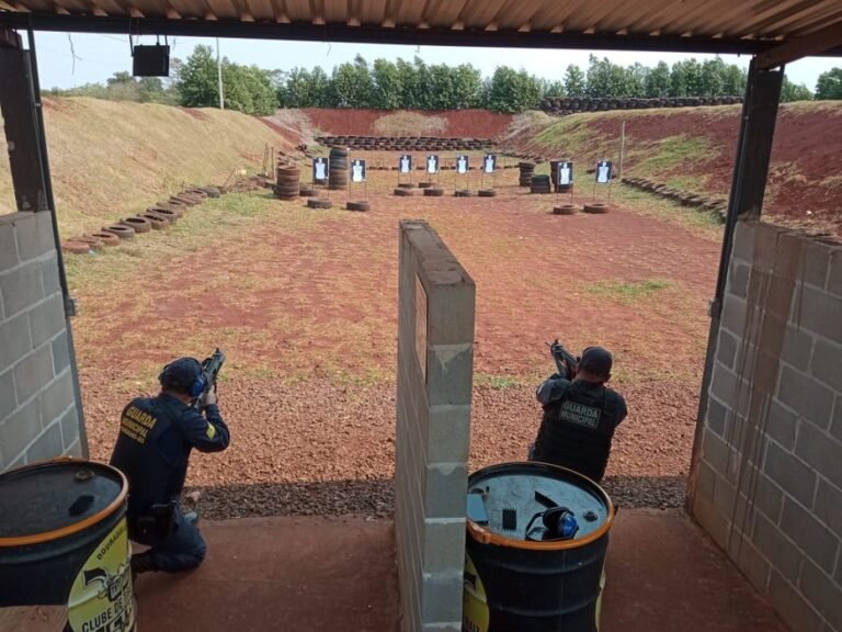 Two officers in a shooting range crouch behind barrels, aiming at paper targets downrange under a covered shelter.