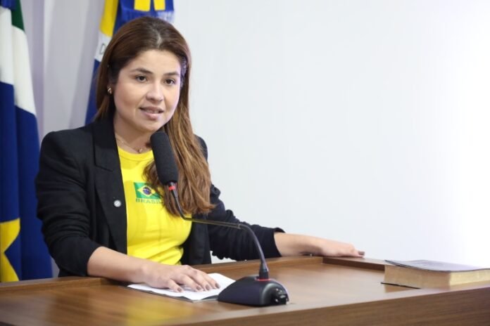 Woman in a yellow shirt and black blazer speaks into a microphone at a wooden podium with papers, flags in the background (official briefing).