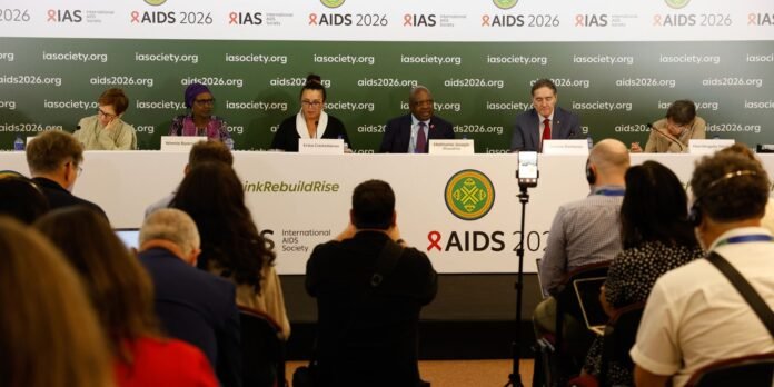 Panel of six experts seated behind a long white table on a stage, speaking into microphones at the AIDS 2026 conference, with a green backdrop and IAS logos behind them.