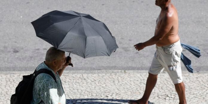 Older man in a patterned shirt walks on a cobblestone sidewalk holding a black umbrella, while a shirtless man passes by in shorts.